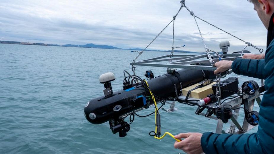 A robot for seagrass seed dispersal hangs by a crane over the ocean surface.